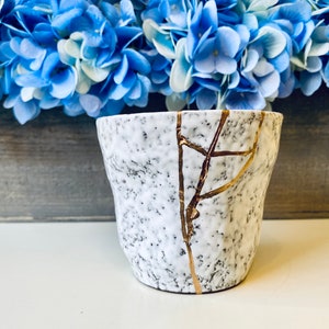Pode incluir: Vaso de cer&acirc;mica branco com superf&iacute;cie texturizada e design de rachaduras douradas. O vaso est&aacute; sobre uma superf&iacute;cie de madeira.