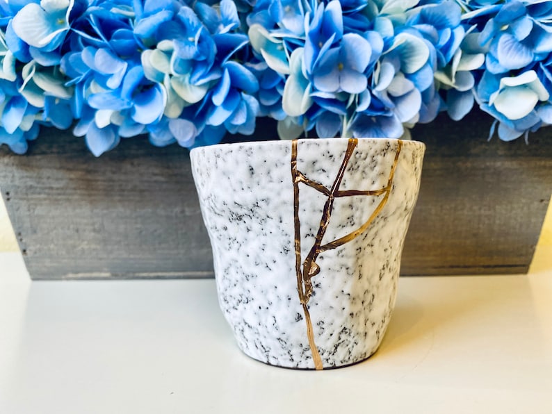 Pode incluir: Vaso de cer&acirc;mica branco com superf&iacute;cie texturizada e design de rachaduras douradas. O vaso est&aacute; sobre uma superf&iacute;cie de madeira.