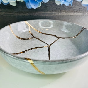 May include: A small, gray ceramic bowl with a crackled glaze and gold accents. The bowl is perfect for holding jewelry or other small items.