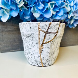 Pode incluir: Uma caneca de cer&acirc;mica branca com esmalte craquelado e detalhes dourados. A caneca tem uma superf&iacute;cie texturizada e &eacute; ligeiramente mais larga na parte superior do que na inferior.