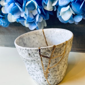 Pode incluir: Caneca de cer&acirc;mica branca com textura pontilhada e rachaduras douradas. A caneca est&aacute; sobre uma superf&iacute;cie branca.