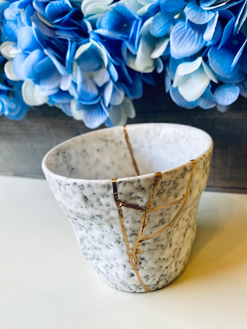 Pode incluir: Caneca de cer&acirc;mica branca com textura pontilhada e rachaduras douradas. A caneca est&aacute; sobre uma superf&iacute;cie branca.