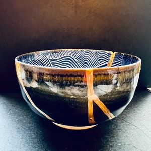 Puede incluir: Un cuenco grande de cerámica negra con un patrón geométrico blanco y dorado. El cuenco tiene una grieta que recorre el lateral, que se destaca con pintura dorada.