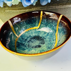 May include: An oval ceramic bowl with a glossy finish. The bowl features a dark brown rim, a teal and green interior, and gold-colored lines. The exterior is a light yellow color. The bowl is a decorative item.