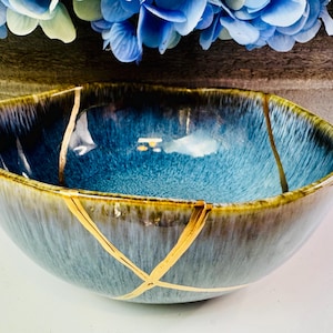 May include: A ceramic bowl featuring a deep blue and teal reactive glaze with a smooth, rounded rim. The exterior and interior surfaces are decorated with prominent gold-colored lines that mimic the appearance of kintsugi repair techniques.