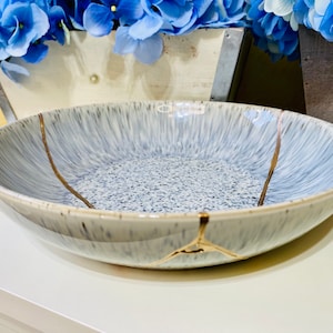 May include: A large, white ceramic bowl with a blue and gray speckled glaze. The bowl has a gold crackle design around the rim and a gold crackle line down the center.