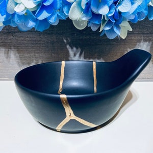 May include: A black ceramic bowl with a gold crackle design and a handle. The bowl is on a white surface.