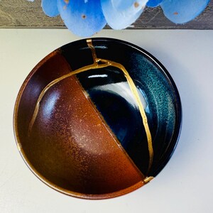 May include: A ceramic bowl with a unique design. The bowl is divided into sections of brown, black, and teal. Gold-colored lines highlight the cracks, creating a striking contrast. The bowl's rim is also gold. This piece is a beautiful example of Kintsugi art.