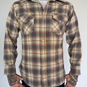 May include: A long-sleeved, button-down shirt with a plaid pattern in shades of beige, brown, and blue. The shirt has two chest pockets with button closures and a pointed collar. The shirt is made of a soft, flannel-like material.