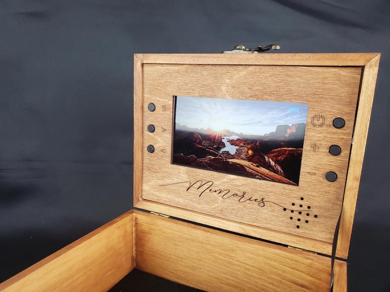 K&ouml;nnte beinhalten: Eine Holzkiste mit einem eingebauten Bildschirm, der ein Landschaftsfoto zeigt. Die Kiste hat einen Klappdeckel und ist mit dem Wort "Erinnerungen" graviert.