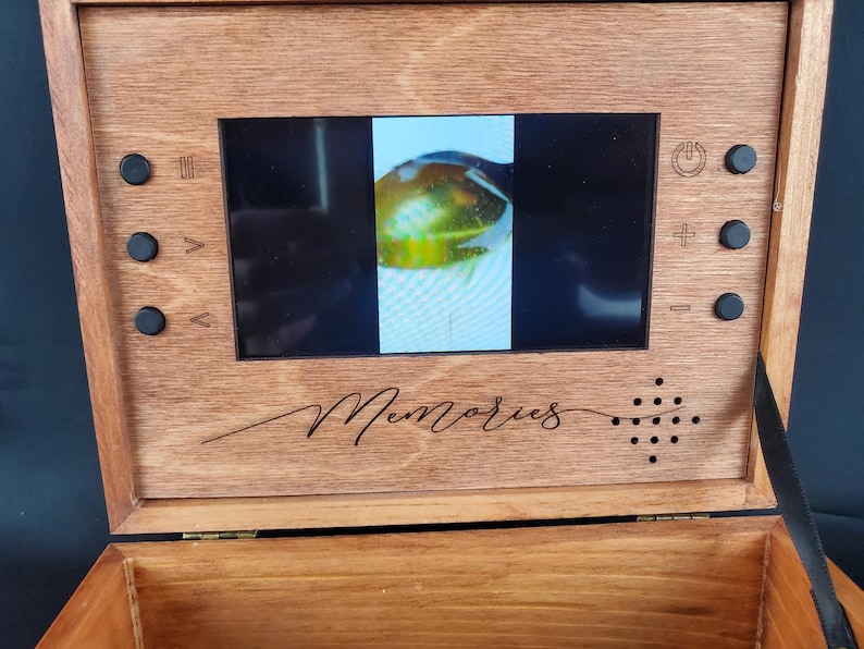 K&ouml;nnte beinhalten: Eine Holzkiste mit einem kleinen Bildschirm, der ein Video von einem gr&uuml;n-gelben Objekt zeigt. Die Kiste ist mit "Memories" beschriftet und hat Kn&ouml;pfe zur Steuerung des Videos.