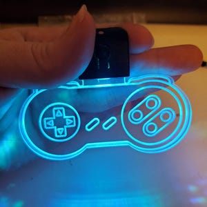 Könnte beinhalten: Ein leuchtender, blauer Acryl-Schlüsselanhänger in Form eines Controllers. Das Design des Controllers umfasst ein Steuerkreuz, Knöpfe und einen rechteckigen oberen Teil. Die Kanten des Controllers sind mit hellem blauem Licht beleuchtet.