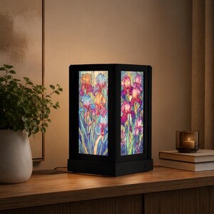 May include: A rectangular table lamp with a black frame and illuminated panels featuring a colorful floral design of irises in shades of purple, blue, orange, and green. The lamp has a solid black base and emits a warm glow through the translucent panels.