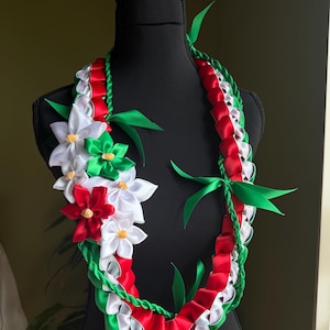 Può includere: Una collana lei decorativa con nastri rossi, bianchi e verdi e fiori di stoffa. La lei è esposta su un manichino nero. I fiori sono bianchi e rossi con centri gialli. Foglie e nastri verdi accentuano il design.