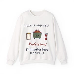 Puede incluir: Una sudadera blanca de cuello redondo con el texto CLAIMS ADJUSTER, Professional Dumpster Fire HANDLER y un gráfico de un portapapeles, un contenedor de basura sonriente en llamas y un extintor rojo en el centro.