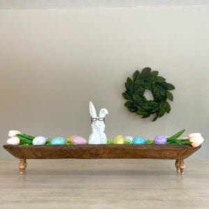 May include: A wooden farmhouse style trough with a white bunny figurine wearing glasses, pink tulips, and pastel colored Easter eggs. The trough is sitting on a wooden table.