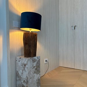 May include: A modern table lamp with a dark blue lampshade and a wooden base. The lamp sits atop a marble-patterned square pedestal. The lamp is plugged into a wall outlet.