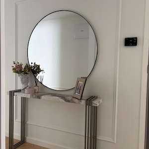 May include: A contemporary hallway arrangement with a circular black-framed mirror above a marble-effect console table. The table has a silver metal frame. There are decorative items, including a framed photo, flowers, and a bust. The wall is white with moulding.