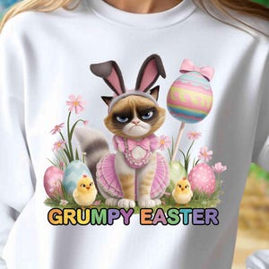 May include: White sweatshirt featuring a grumpy cat wearing bunny ears and a pink dress, surrounded by Easter eggs and chicks. The text "GRUMPY EASTER" is displayed in colorful letters.