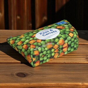 K&ouml;nnte beinhalten: Ein Rei&szlig;verschlussetui mit einem lebendigen Aufdruck von Erbsen und gew&uuml;rfelten Karotten. Das Design enth&auml;lt den Text "Peas & Carrots" auf einem wei&szlig;en Oval, mit einem Nettogewicht von 340g. Das Etui befindet sich vor einem Holzgrund.