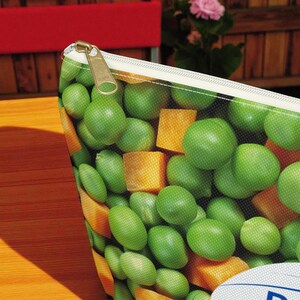 K&ouml;nnte beinhalten: Ein Rei&szlig;verschlussetui mit einem lebendigen Aufdruck aus gr&uuml;nen Erbsen und orangefarbenen gew&uuml;rfelten Karotten. Das Etui hat einen wei&szlig;en Rei&szlig;verschluss und einen goldfarbenen Rei&szlig;verschlussanh&auml;nger. Der Hintergrund ist ein Holztisch.