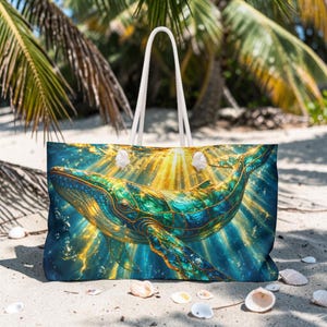 Könnte beinhalten: Eine große Strandtasche mit einer lebendigen Illustration eines Wals in Blau-, Grün- und Goldtönen. Der Wal ist vor einem Hintergrund aus strahlendem Licht und Blasen platziert. Die Tasche hat weiße Seilgriffe.