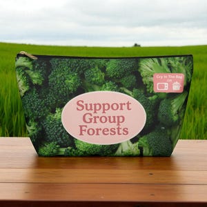 Könnte beinhalten: Eine Reißverschlusstasche mit einem Muster aus grünen Brokkoli-Röschen. Die Tasche hat ein hellrosa Oval mit den Worten "Support Group Forests" und ein kleines rosa Quadrat mit Kochanweisungen. Der Hintergrund ist ein grünes Feld.