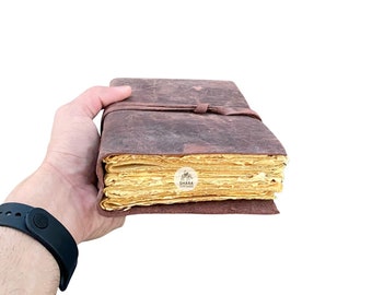 May include: A brown leather-bound journal with yellowed pages. The journal is closed and tied with a brown leather strap. The journal is held in a hand.