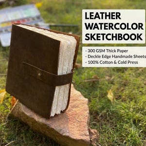 May include: A brown leather sketchbook with a strap and brass button closure, featuring thick white deckle edge handmade paper. The text reads LEATHER WATERCOLOR SKETCHBOOK, 300 GSM Thick Paper, Deckle Edge Handmade Sheets, 100% Cotton & Cold Press.