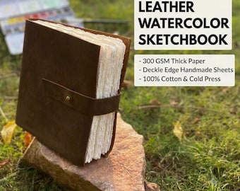May include: A brown leather sketchbook with a strap and brass button closure, featuring thick white deckle edge handmade paper. The text reads LEATHER WATERCOLOR SKETCHBOOK, 300 GSM Thick Paper, Deckle Edge Handmade Sheets, 100% Cotton & Cold Press.