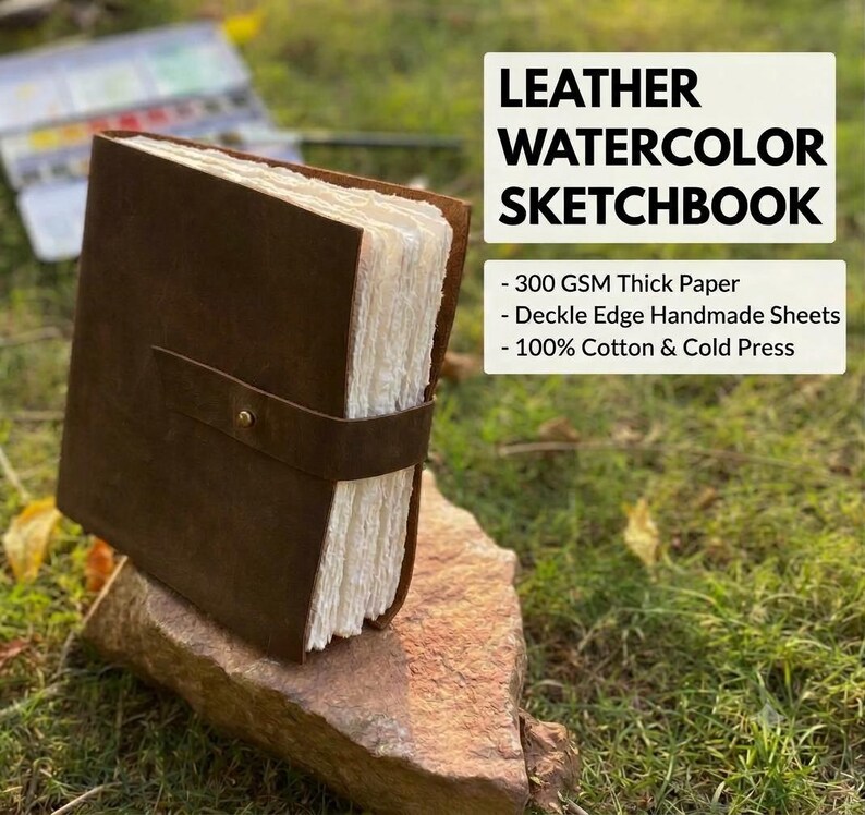 May include: A brown leather sketchbook with a strap and brass button closure, featuring thick white deckle edge handmade paper. The text reads LEATHER WATERCOLOR SKETCHBOOK, 300 GSM Thick Paper, Deckle Edge Handmade Sheets, 100% Cotton & Cold Press.