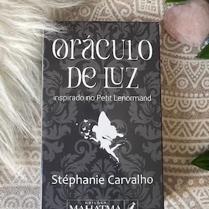 Puede incluir: Un libro titulado "Oráculo De Luz" con una cubierta negra con una ilustración de hada blanca y texto. El libro está inspirado en Petit Lenormand y escrito por Stéphanie Carvalho. El libro está sobre una superficie texturizada.