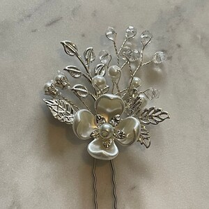 May include: A silver hair comb with a white flower and crystal accents. The comb has two prongs for easy insertion into hair.