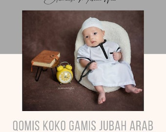 May include: A baby wearing a white Qomis Koko Gamis Jubah Arab outfit with black trim, a white cap, and a string of beads. The image includes a yellow alarm clock, a small brown book, and the text "Atthalifi Moslem Wear".