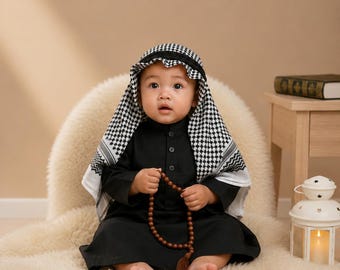 Puede incluir: Una túnica negra de manga larga usada con un pañuelo keffiyeh de cuadros blancos y negros. La prenda presenta un cuello abotonado y se combina con un rosario de cuentas marrones sostenido en las manos.