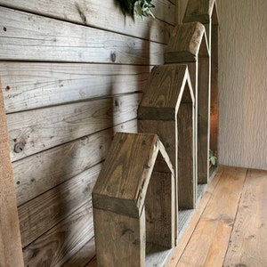 K&ouml;nnte beinhalten: Ein Satz aus vier Holzregalen in Hausform, in absteigender Gr&ouml;&szlig;e gestapelt, mit einem rustikalen grauen Finish. Die Regale sind aus wiederverwendetem Holz gefertigt und haben ein nat&uuml;rliches, verwittertes Aussehen.