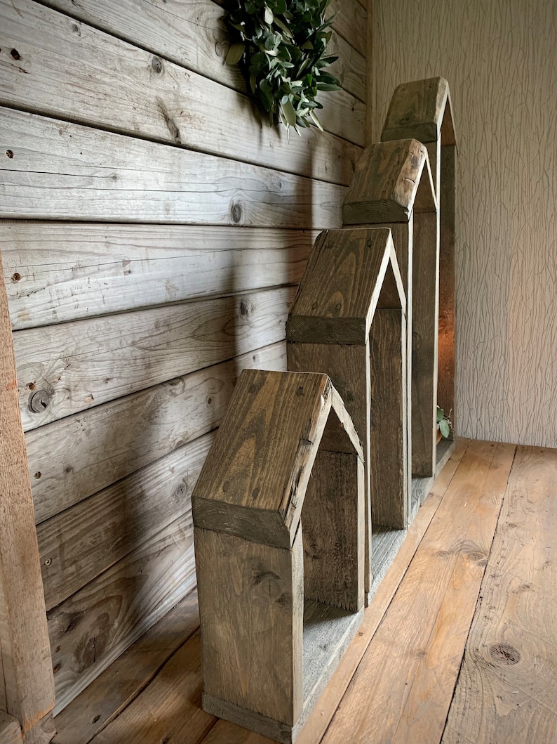 K&ouml;nnte beinhalten: Ein Satz aus vier Holzregalen in Hausform, in absteigender Gr&ouml;&szlig;e gestapelt, mit einem rustikalen grauen Finish. Die Regale sind aus wiederverwendetem Holz gefertigt und haben ein nat&uuml;rliches, verwittertes Aussehen.
