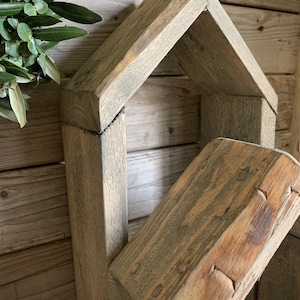 K&ouml;nnte beinhalten: Ein rustikales, hausf&ouml;rmiges Holzregal mit einer verwitterten grauen Oberfl&auml;che. Das Regal hat eine rechteckige &Ouml;ffnung und ein schr&auml;ges Dach. Das Holz ist rau und hat eine nat&uuml;rliche Maserung.