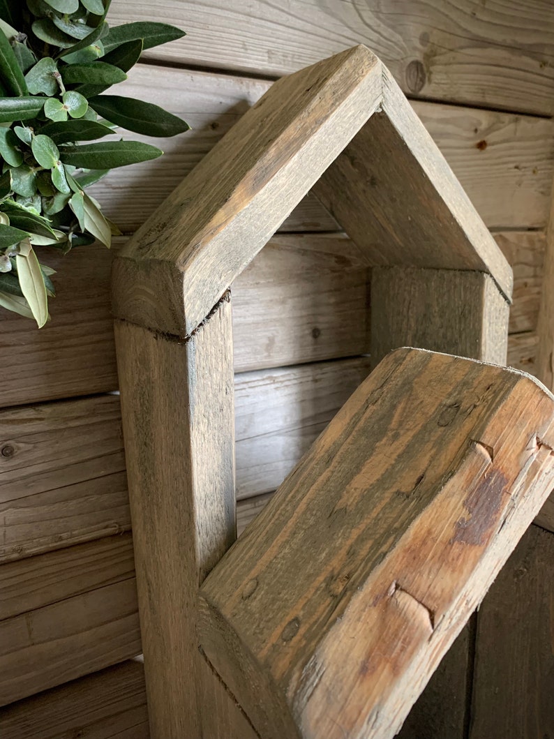 K&ouml;nnte beinhalten: Ein rustikales, hausf&ouml;rmiges Holzregal mit einer verwitterten grauen Oberfl&auml;che. Das Regal hat eine rechteckige &Ouml;ffnung und ein schr&auml;ges Dach. Das Holz ist rau und hat eine nat&uuml;rliche Maserung.