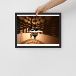 May include: A framed photograph of a decorative wall lamp. The lamp is black with a perforated shade, casting a patterned light on the wall. The frame is black, and the image is held by a hand.