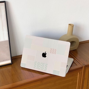 Puede incluir: Un ordenador portátil con una funda estampada con un diseño de patchwork en colores pastel. La funda tiene varios patrones geométricos, incluyendo rayas y zigzags. El logotipo de Apple es visible. El portátil está sobre una superficie de madera.