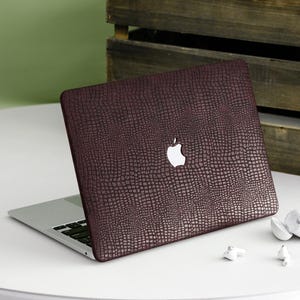Könnte beinhalten: Eine weinrote Laptop-Hülle mit einem strukturierten, reptilienartigen Muster. Die Hülle befindet sich auf einem silbernen Laptop mit einem weißen Apple-Logo. Weiße kabellose Ohrhörer liegen neben dem Laptop.