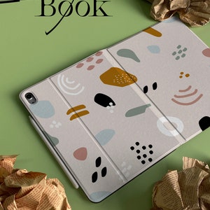 Könnte beinhalten: Eine hellgraue Tablethülle mit einem geometrischen Muster in Braun-, Rosa-, Blau- und Weißtönen. Die Hülle hat eine Klappe, die sich über das Tablet legt. Das Wort "Book" ist in Schwarz auf grünem Hintergrund geschrieben.