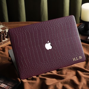 May include: A deep burgundy laptop case with a crocodile skin effect. The case displays the Apple logo and gold initials "H.L.B." The laptop is open, showing the screen. A chess set and candle are in the background.