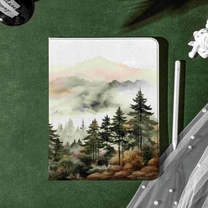 Puede incluir: Una pintura de acuarela de un paisaje montañoso brumoso con un bosque de pinos en primer plano. La pintura está sobre un fondo blanco y está enmarcada en un marco blanco.