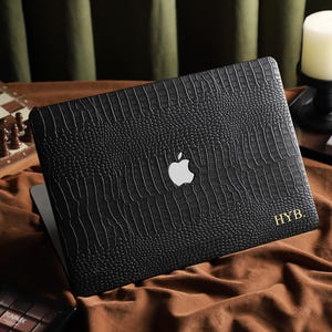 May include: Black laptop case with a textured, crocodile-like pattern. The Apple logo is visible, along with the initials "HYB" in gold. The case is on a brown surface, with a chess set and candle in the background.