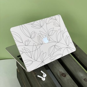 Puede incluir: Un portátil blanco con un diseño floral negro en la tapa. El portátil está cerrado y sentado en una caja de madera. Hay auriculares inalámbricos blancos en la caja.