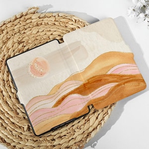 Könnte beinhalten: Eine Aquarell-Illustration einer Landschaft mit rosa, braunen und goldenen Hügeln und einer großen, blassrosa Sonne. Die Illustration ist auf einem schwarzen Umschlag für ein Notizbuch oder Tagebuch gedruckt.