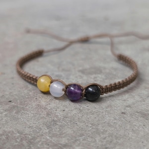 Könnte beinhalten: Ein Makramee-Armband mit einer Reihe farbiger Edelsteinperlen. Das Armband hat eine beige Kordel und enthält gelbe, weiße, lila und schwarze Perlen. Das Armband ist verstellbar.