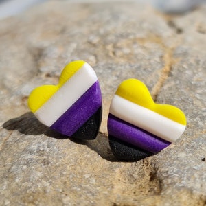 Könnte beinhalten: Ein Paar herzförmige Ohrringe in den Farben der Non-Binary-Pride-Flagge. Die Ohrringe sind gelb, weiß, lila und schwarz.